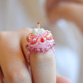 Pink beaded cake ring with a small decorative item on a finger against a blurred background