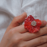 Hand holding a red beaded flower ring with a silver center on a white fabric background.
