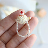 beaded ring with a cupcake design held on a hand against a blurred background