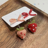 Beaded strawberry and bow-shaped earrings on a wooden surface with a white card.