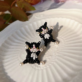 Black beaded cat earrings on a white shell with blurred floral background