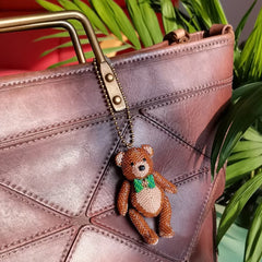 Brown teddy bear bag charm attached to a leather bag with green plants in the background