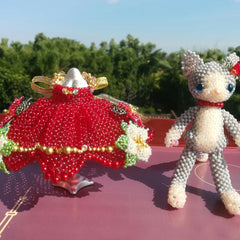 Two decorative figurines, one red wedding dress, the other beaded cat, on a pink surface with greenery in the background.