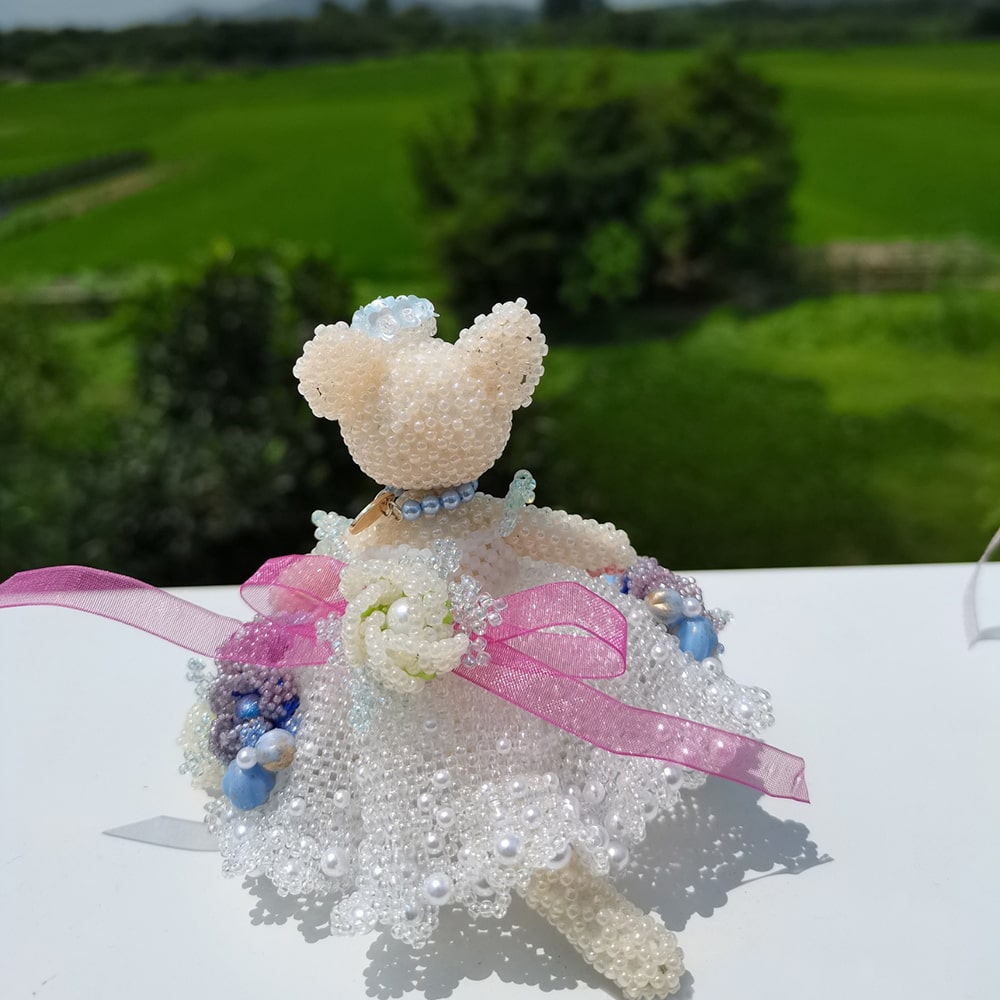handmade beaded cat in a white wedding dress with pink ribbons on a table outdoors.