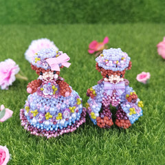 Two handmade bear figurines made of beads on a grassy background with pink flowers.