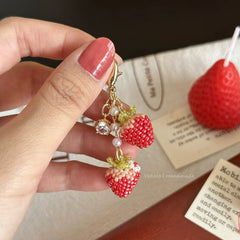 Hand holding strawberry-shaped bag chams with a background of a red strawberry and a card.