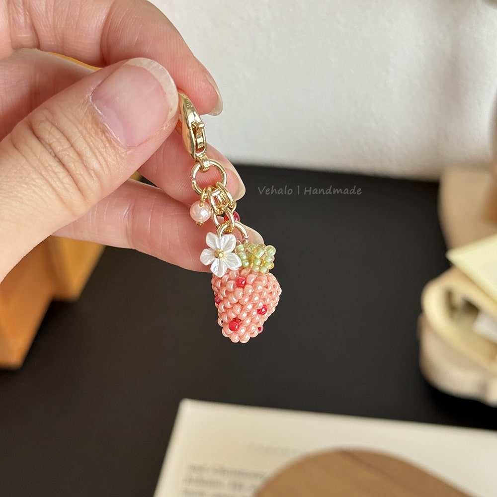 Hand holding a strawberry-shaped keychain with beads and a flower charm on a dark surface.