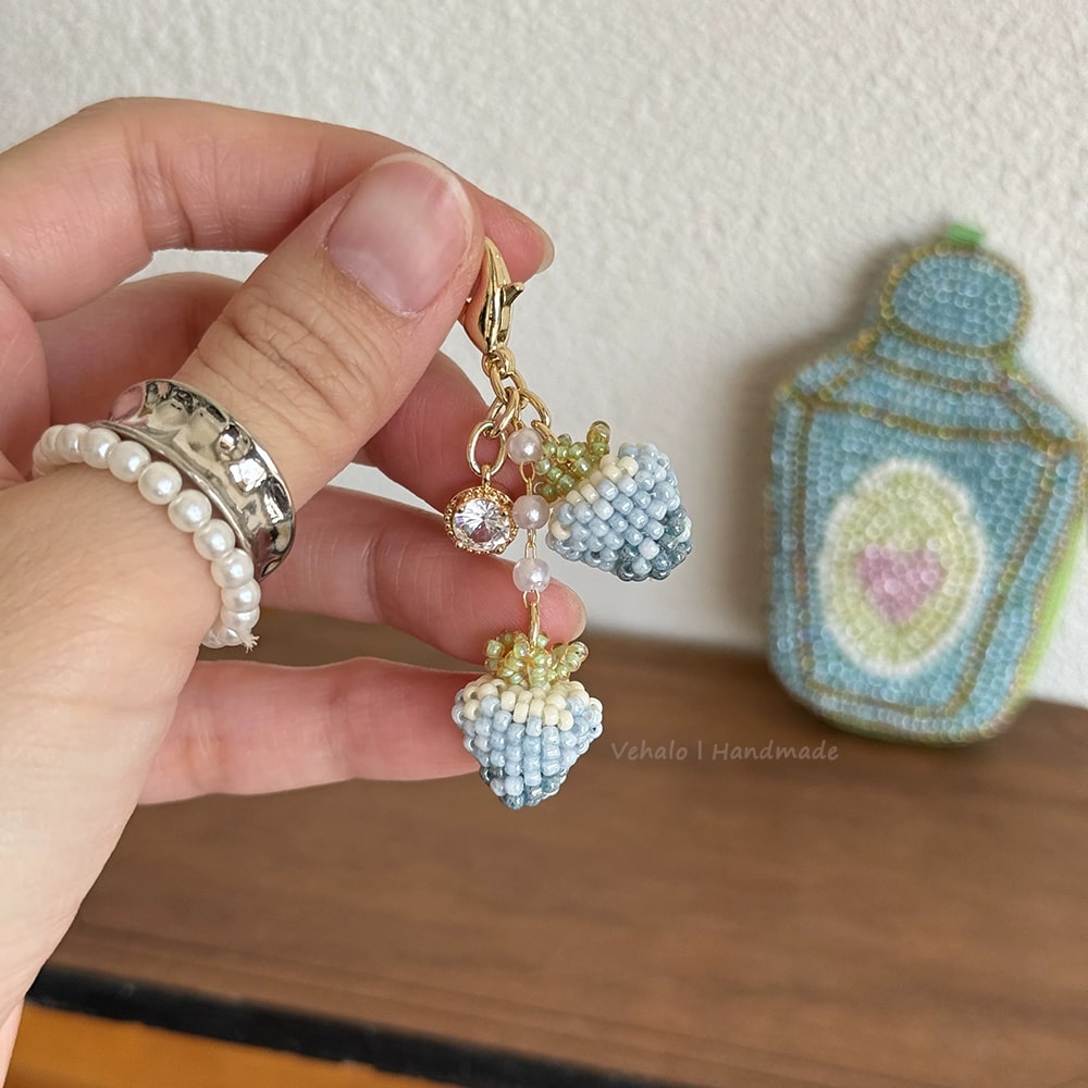 Hand holding a colorful keychain with beads and charms, with a needlepoint item in the background.