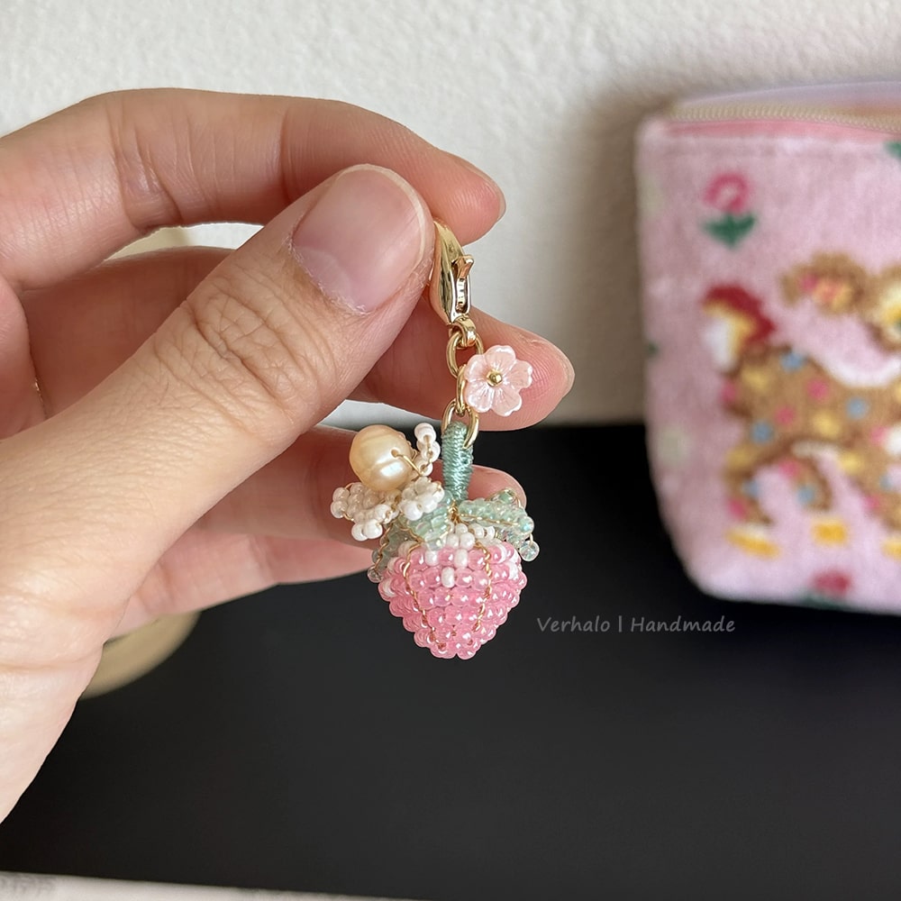 Hand holding a decorative strawberry keychain with beads and flowers, with a colorful pouch in the background.