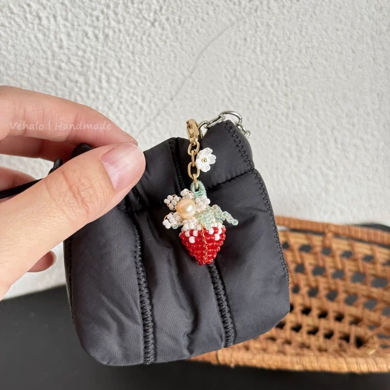 Hand holding a small black pouch with a decorative strawberry keychain against a neutral background.