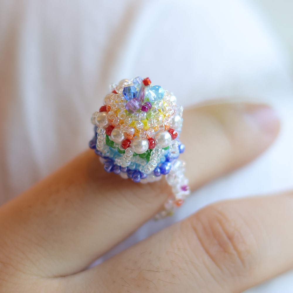Colorful beaded rainbow cake ring on a finger with a blurred background