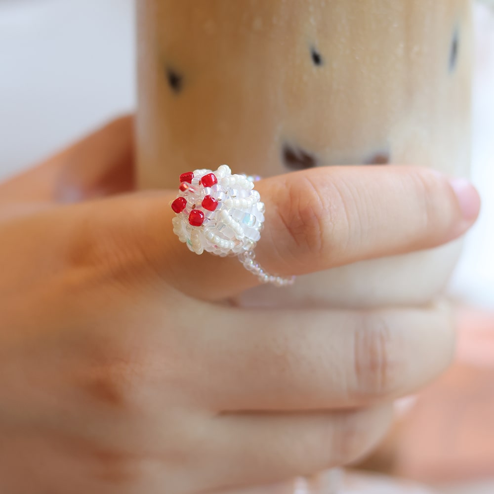 Close-up of a hand wearing a white beaded ring with red beads, holding a blurred cup.