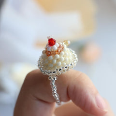 Decorative pudding ring with pearls and red gemstone held by a hand against a blurred background