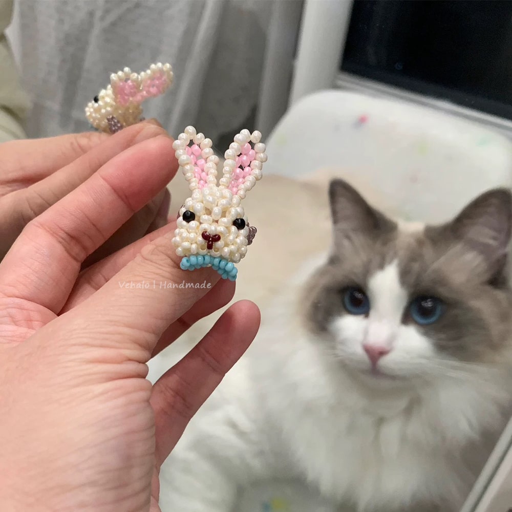 Handmade beaded bunny ring held by a hand with a cat in the background