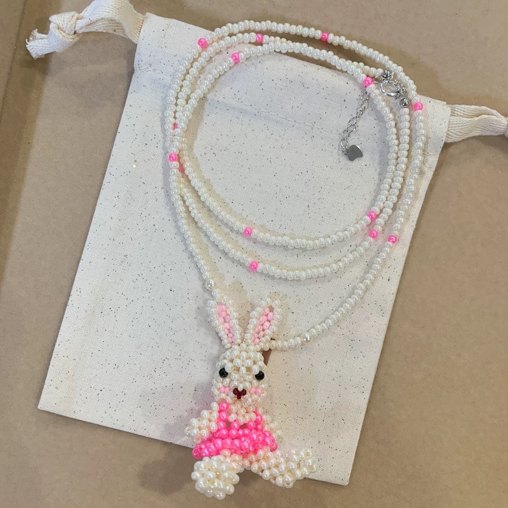 Beaded bunny necklace with pink dress on a white drawstring bag against a beige background