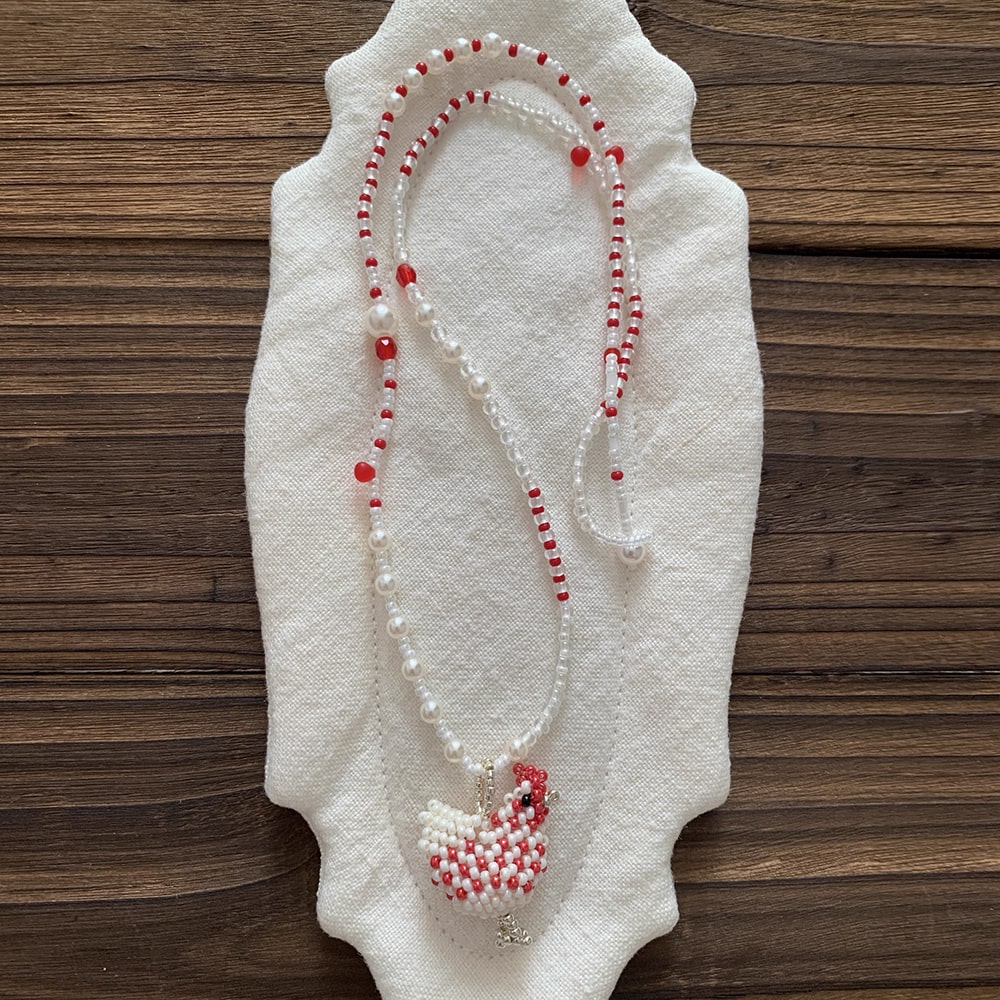 Beaded necklace with red and white chcicken pendant on a white fabric background - Verhalo Jewelry