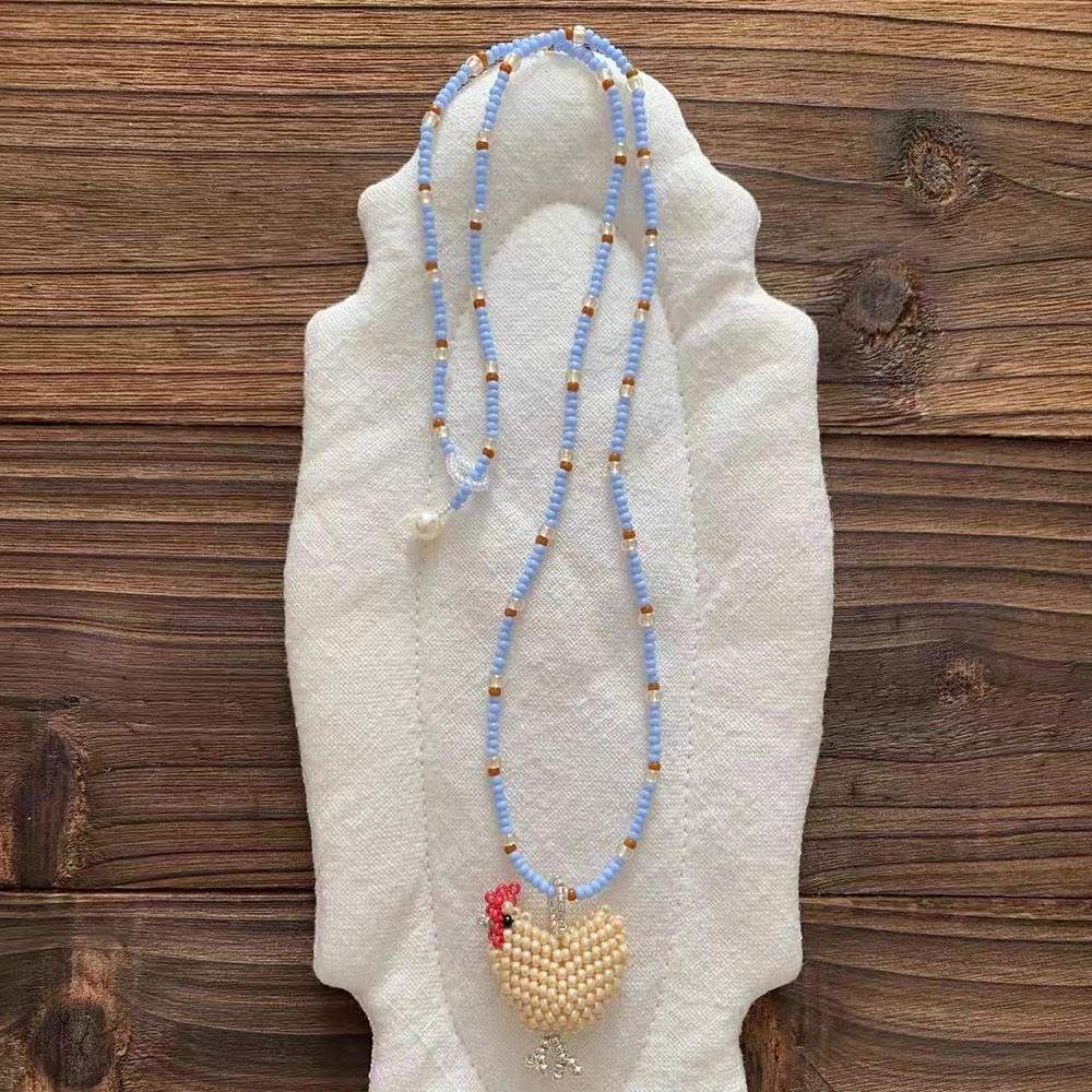 Beaded necklace with a white chicken charm on a mannequin hand against a wooden background - Verhalo Jewelry