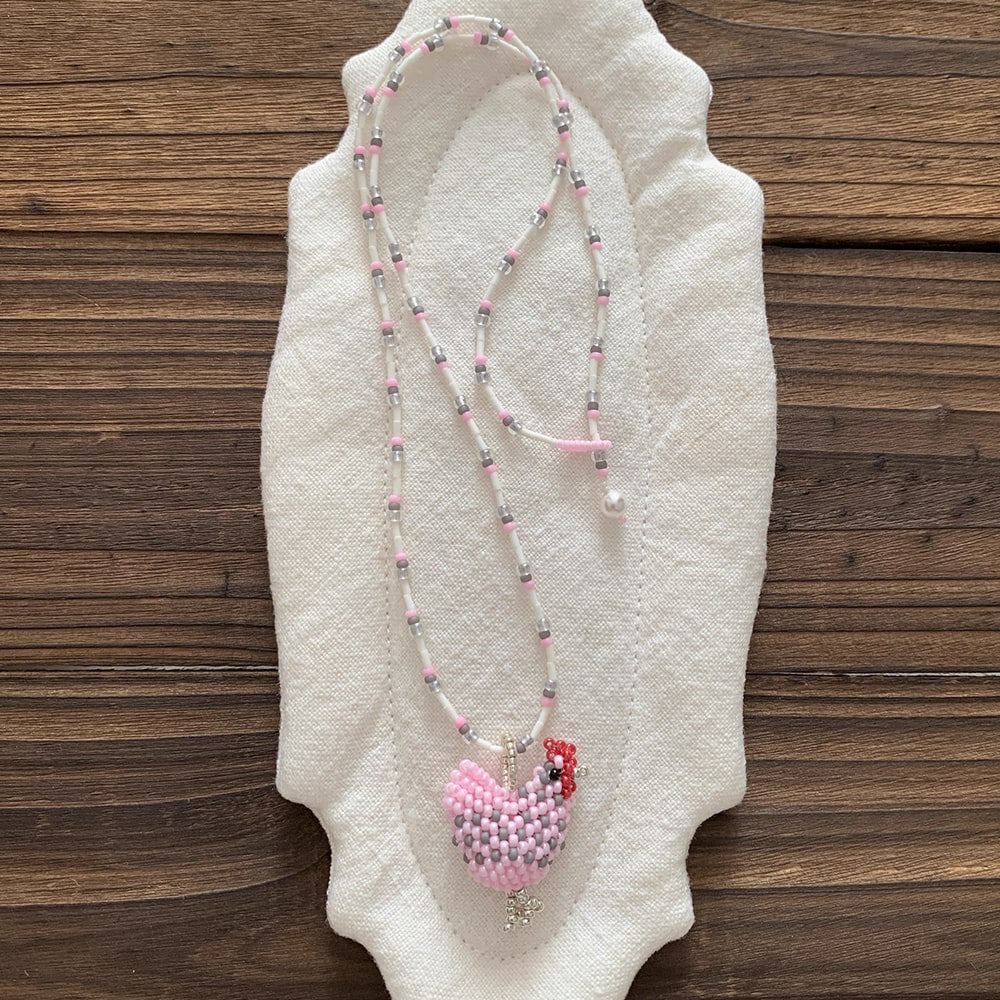 Beaded necklace with a pink chicken pendant on a textured white surface with a wooden background - Verhalo Jewelry