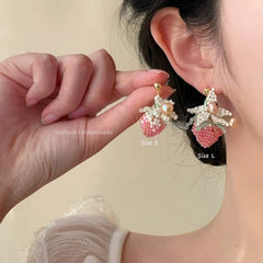 Person wearing strawberry-shaped earrings with pearl details, held by a hand against a neutral background.